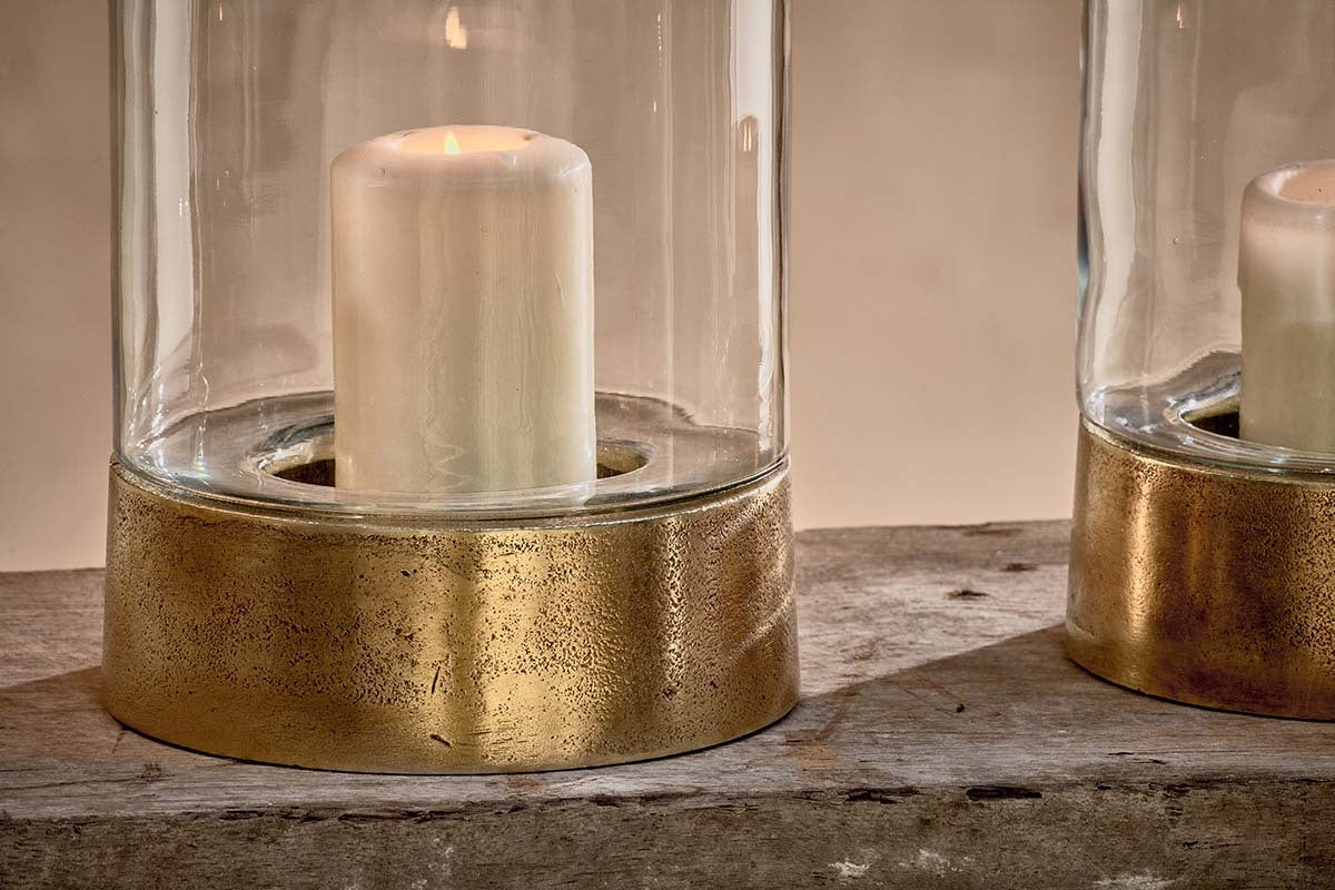 The Rajura Hurricane Lantern by nkuku features a white pillar candle in a cylindrical glass with a textured brass-toned base, set on rustic wood. Its warm glow creates an inviting atmosphere, with another Rajura lantern partially visible to the right.