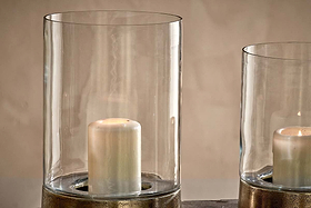 Two white pillar candles glow in nkuku Rajura Hurricane Lanterns—clear cylindrical holders with metallic bases—casting ambient light against a beige backdrop. Set side by side, one lantern is mostly out of frame to the right.