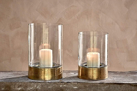 Two Rajura Hurricane Lanterns by nkuku, featuring white pillar candles and metallic gold bases, sit on a rustic wooden surface against a beige wall. The lanterns are different heights, ideal for adding ambient light to any room.