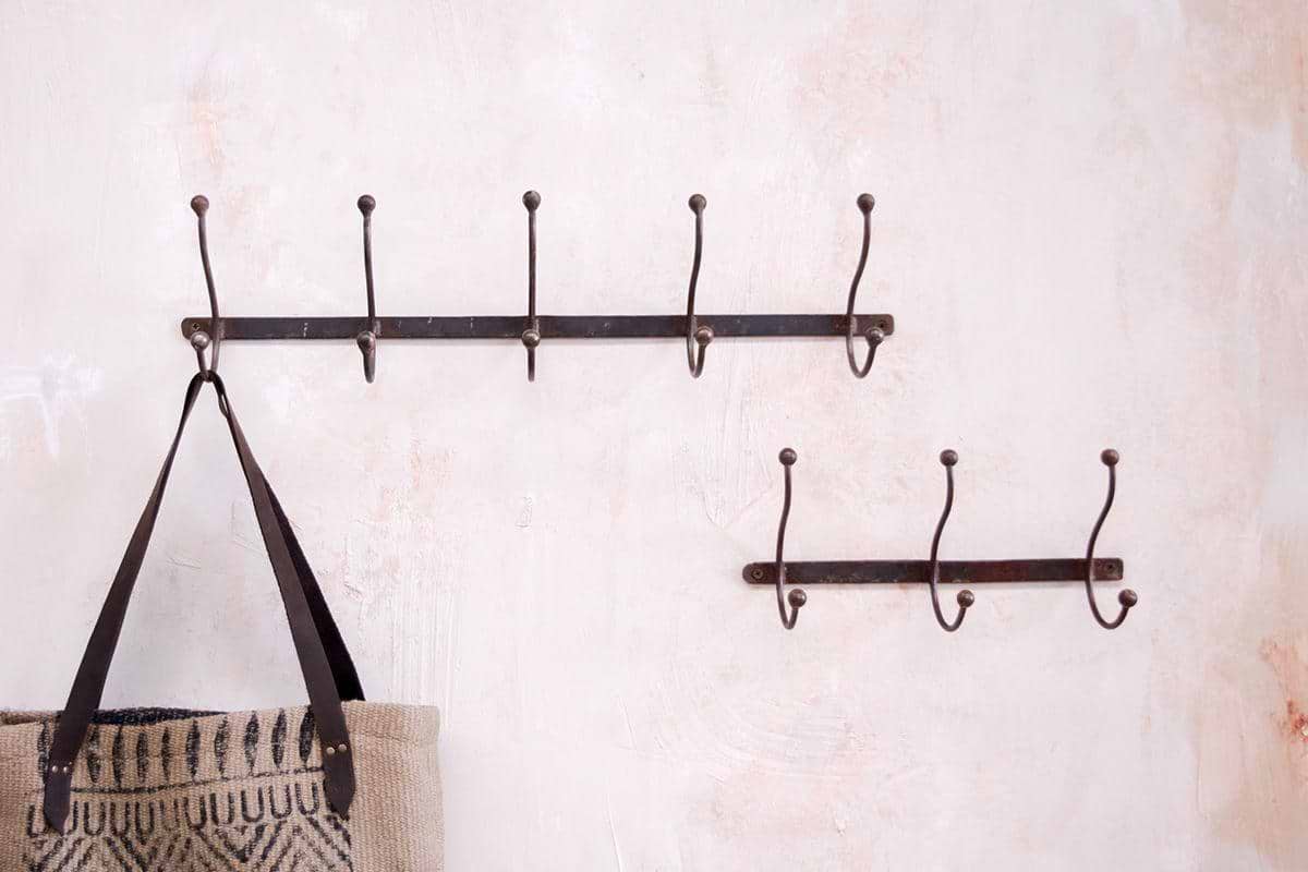 Two black nkuku Rama Iron Hook coat racks are mounted on a textured, pale pink hallway wall. The larger rack holds a beige woven bag with dark handles and geometric pattern; the smaller, below it, is empty—a stylish storage solution.