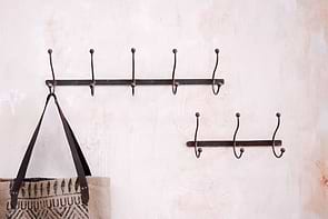 Two black nkuku Rama Iron Hook coat racks are mounted on a textured, pale pink hallway wall. The larger rack holds a beige woven bag with dark handles and geometric pattern; the smaller, below it, is empty—a stylish storage solution.