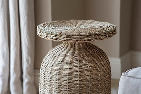 A close-up of the nkuku Rampura Rattan Side Table - Natural highlights its intricate, handwoven texture. Set indoors by light curtains and a neutral wall, it adds a cozy, rustic touch to the space.
