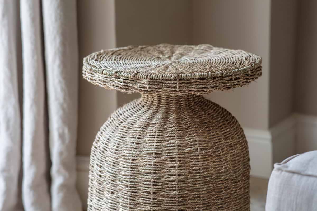 A close-up of the nkuku Rampura Rattan Side Table - Natural highlights its intricate, handwoven texture. Set indoors by light curtains and a neutral wall, it adds a cozy, rustic touch to the space.