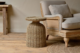 The nkuku Rampura Rattan Side Table - Natural sits beside a beige upholstered armchair with wooden frame. Light wood floors, a round woven rug, and neutral-toned walls add to the cozy, rustic feel.