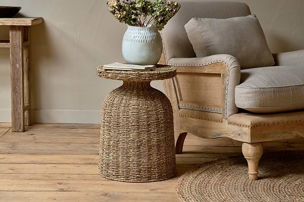 The cozy, rustic living room features light wood floors, a braided rug, and neutral-toned décor. A nkuku Rampura Rattan Side Table – Natural holds a pale ceramic vase with dried flowers beside a beige cushioned wooden armchair.