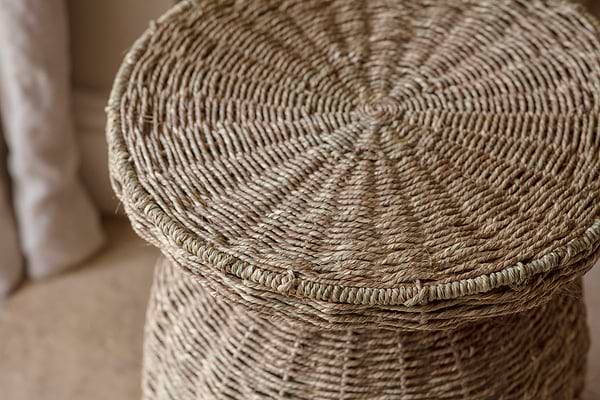 Close-up of the nkuku Rampura Rattan Side Table in Natural, featuring handwoven beige fibers in a tight spiral pattern reminiscent of textured wicker furniture. A curtain and neutral floor set a warm, inviting scene.