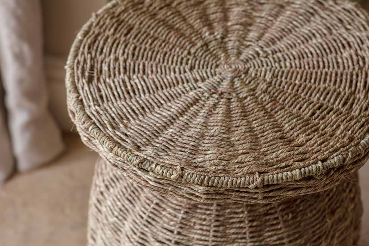 Close-up of the nkuku Rampura Rattan Side Table in Natural, featuring handwoven beige fibers in a tight spiral pattern reminiscent of textured wicker furniture. A curtain and neutral floor set a warm, inviting scene.