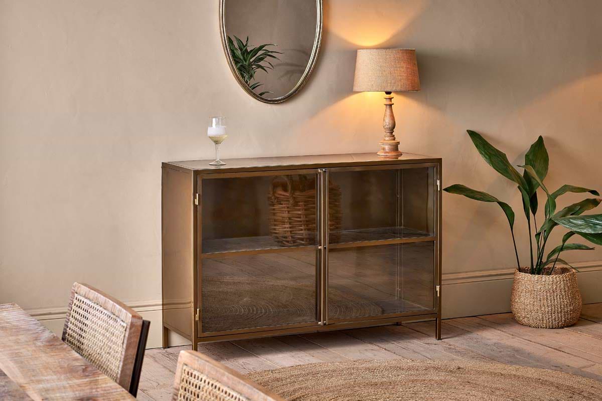 A cozy room features the nkuku Ranchi Iron Sideboard with glass doors, topped with a lamp and drink. A round mirror above reflects a leafy plant, complemented by woven furniture, a round rug, and a tall potted plant.