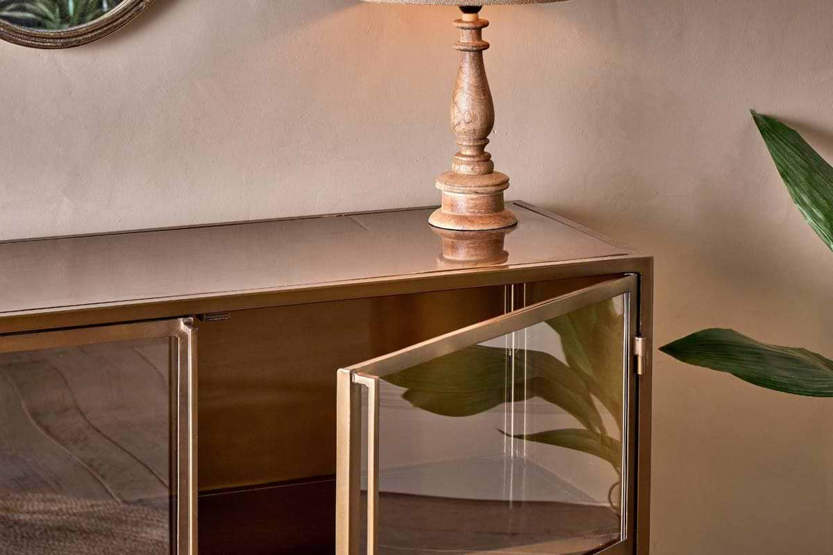 A wooden lamp with a beige shade rests on the nkuku Ranchi Iron Sideboard, featuring glass doors—one partially open. Green leaves are nearby, and the warm, neutral decor complements the scene.
