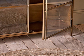 A close-up, eye-level shot features the nkuku Ranchi Iron Sideboard with glass doors—one ajar to reveal a shelf—standing on a distressed wooden floor with a round jute rug in the foreground.