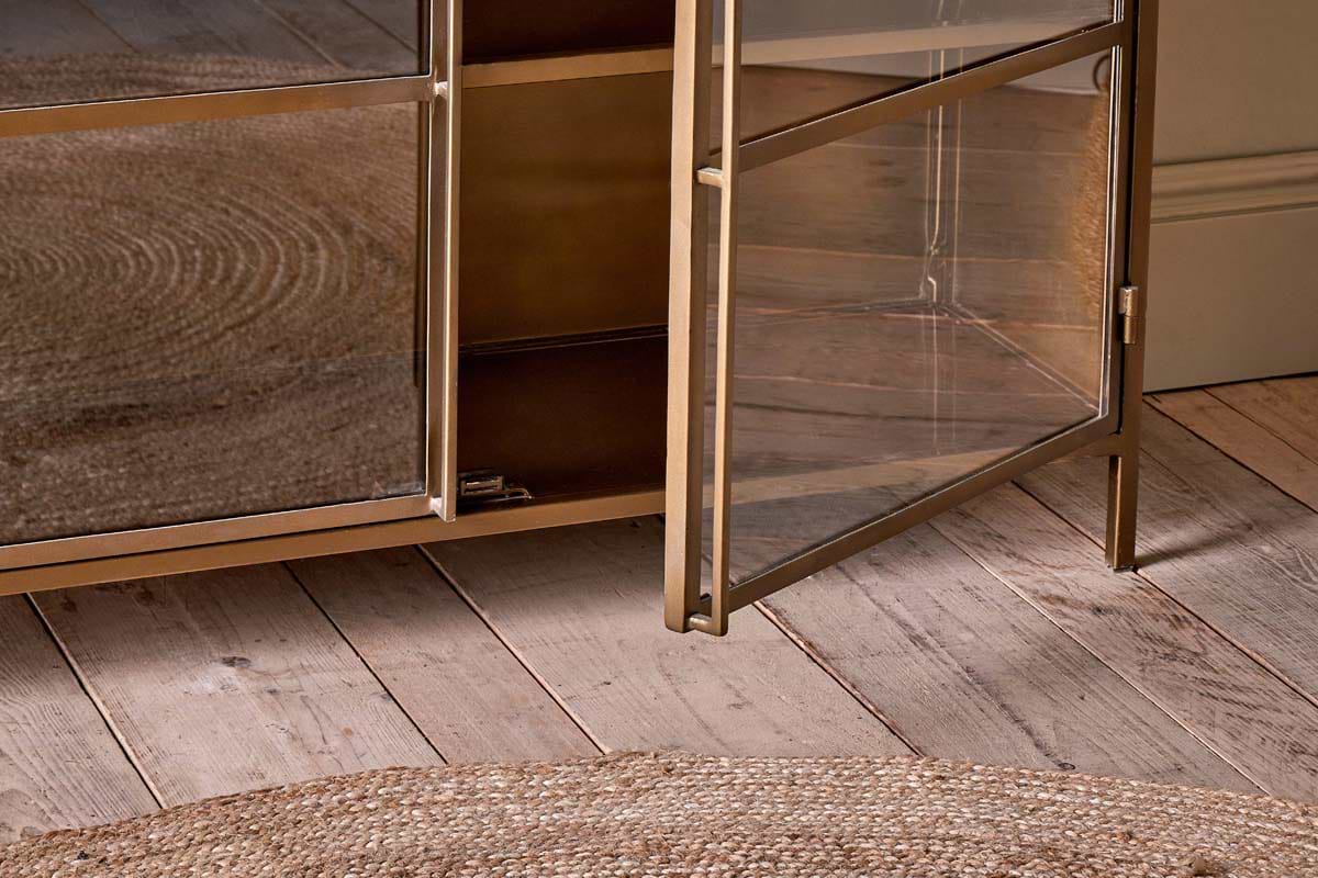 A close-up, eye-level shot features the nkuku Ranchi Iron Sideboard with glass doors—one ajar to reveal a shelf—standing on a distressed wooden floor with a round jute rug in the foreground.