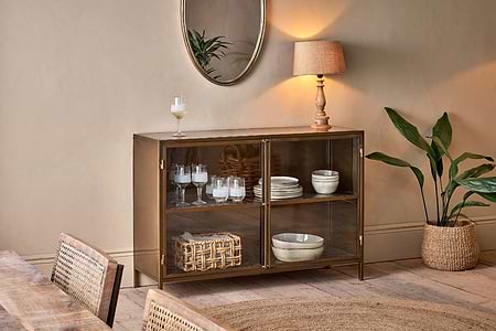 A cozy dining room showcases the nkuku Ranchi Iron Sideboard with glass-fronted doors, displaying tableware and a woven basket. A lamp and cocktail glass rest on top, while a round mirror and green plant accent beige walls and wood floors.