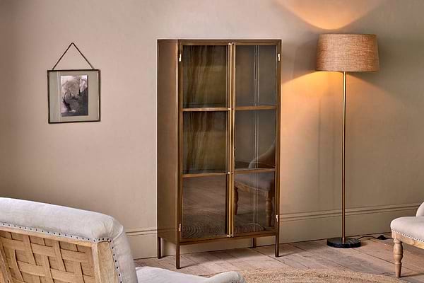 A cozy living room with a beige wall, glass-fronted nkuku Ranchi Iron Tall Cabinet on slender brass legs, cream armchair, floor lamp with beige shade casting a warm glow, and framed art above the partially visible carpet.