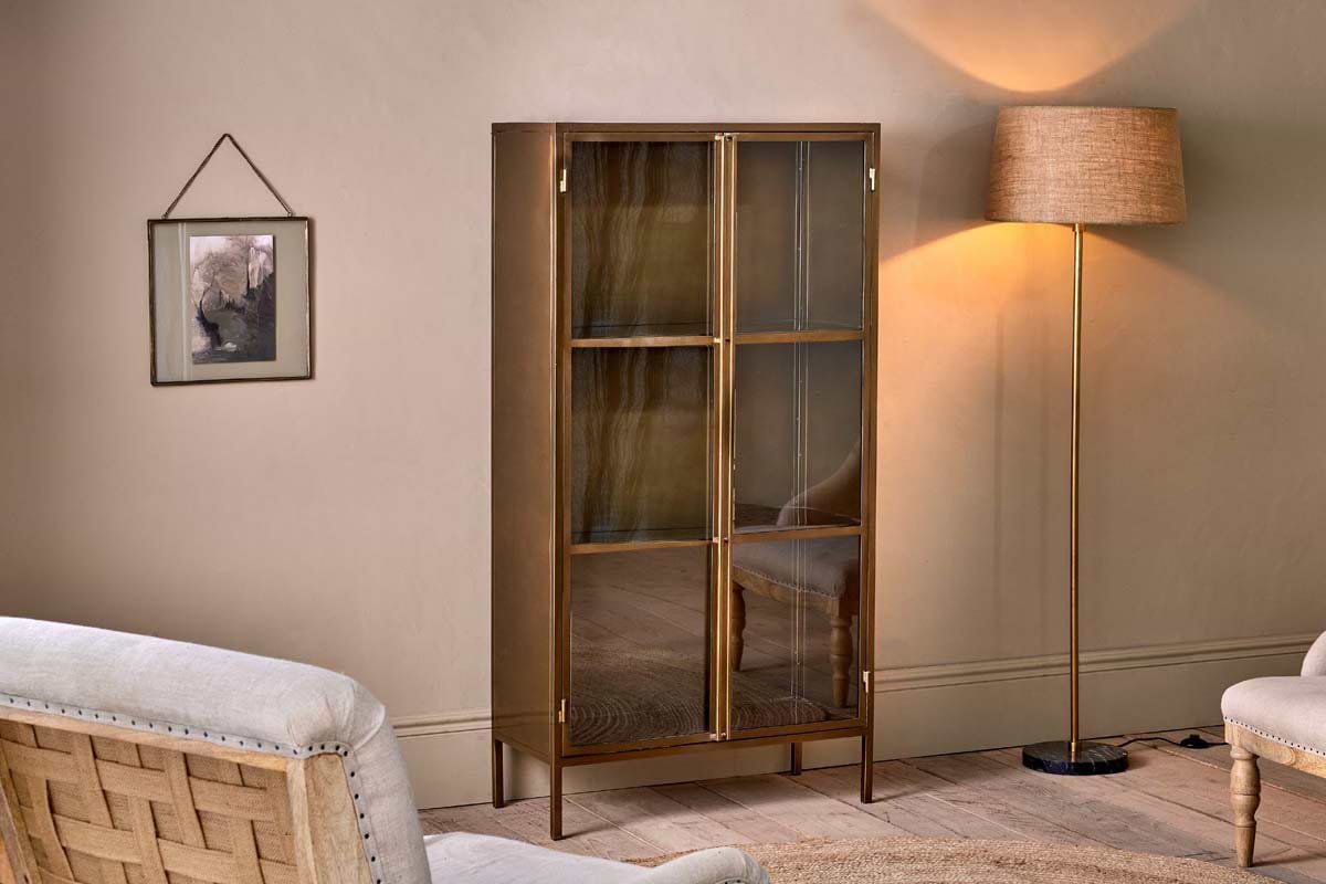 A cozy living room with a beige wall, glass-fronted nkuku Ranchi Iron Tall Cabinet on slender brass legs, cream armchair, floor lamp with beige shade casting a warm glow, and framed art above the partially visible carpet.