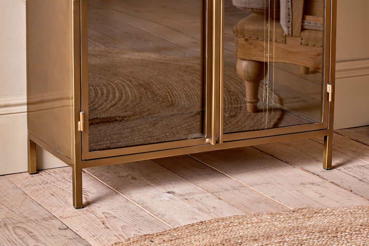 The Ranchi Iron Tall Cabinet by nkuku, featuring gold-framed glass doors, stands on light wood flooring beside a round woven rug, reflecting an upholstered wooden chair and the rug’s pattern against a cream wall and baseboard.