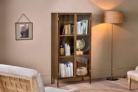 The nkuku Ranchi Iron Tall Cabinet with glass fronted doors displays books, a globe, and decor next to a floor lamp. A beige armchair and woven chair complete the warmly lit room, with a framed picture on the cream wall for coziness.