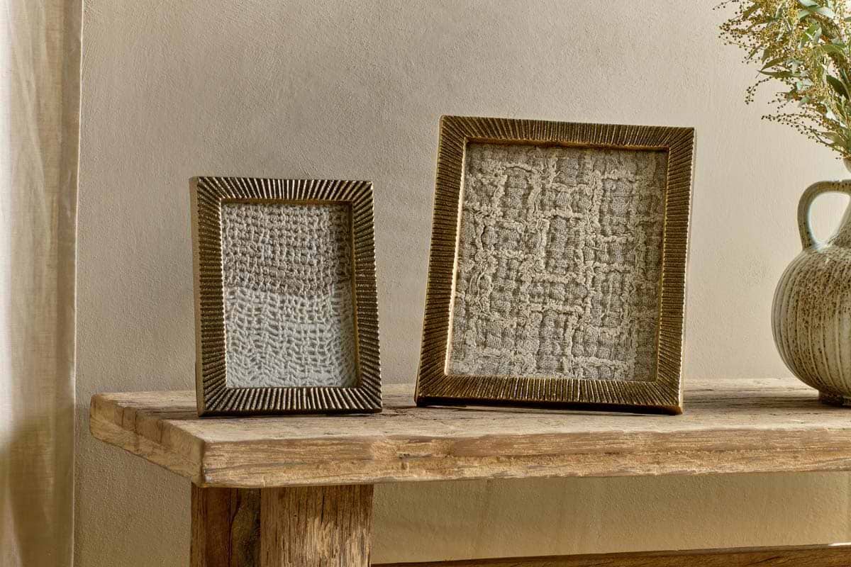Two textured picture frames—a nkuku Ranoli Iron Etched Photo Frame in Antique Brass and a vintage-inspired frame—sit on a rustic wooden table. The frames display abstract, woven white fabric; one is smaller and upright, while the other is larger and square. To the right, a ceramic vase filled with dried flowers completes the arrangement against a light beige wall.