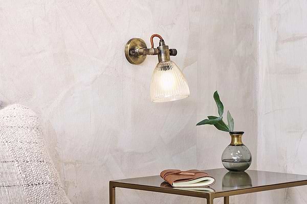 The nkuku Rarni Recycled Glass Single Wall Light casts a warm glow above a small metallic table, which holds a green glass vase with leafy branch, a closed book, and a folded brown leather item for a cozy minimalist look.