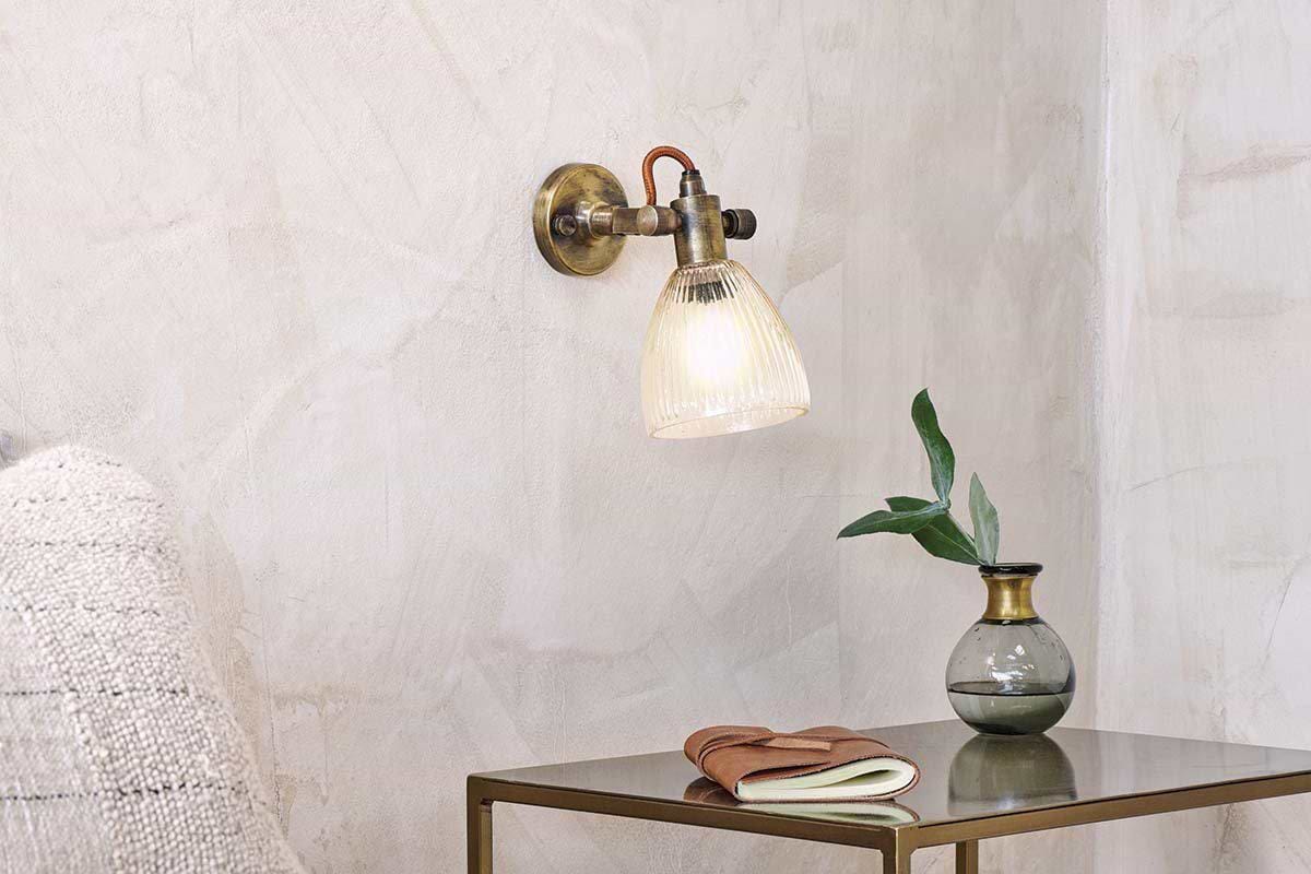 The nkuku Rarni Recycled Glass Single Wall Light casts a warm glow above a small metallic table, which holds a green glass vase with leafy branch, a closed book, and a folded brown leather item for a cozy minimalist look.