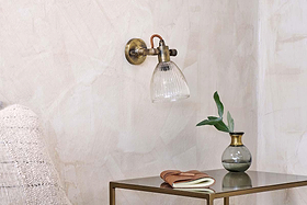 The nkuku Rarni Recycled Glass Single Wall Light is mounted on a textured beige wall above a metal side table with a green-leaf vase, closed notebook, and brown eyeglasses. The edge of a textured chair appears on the left.