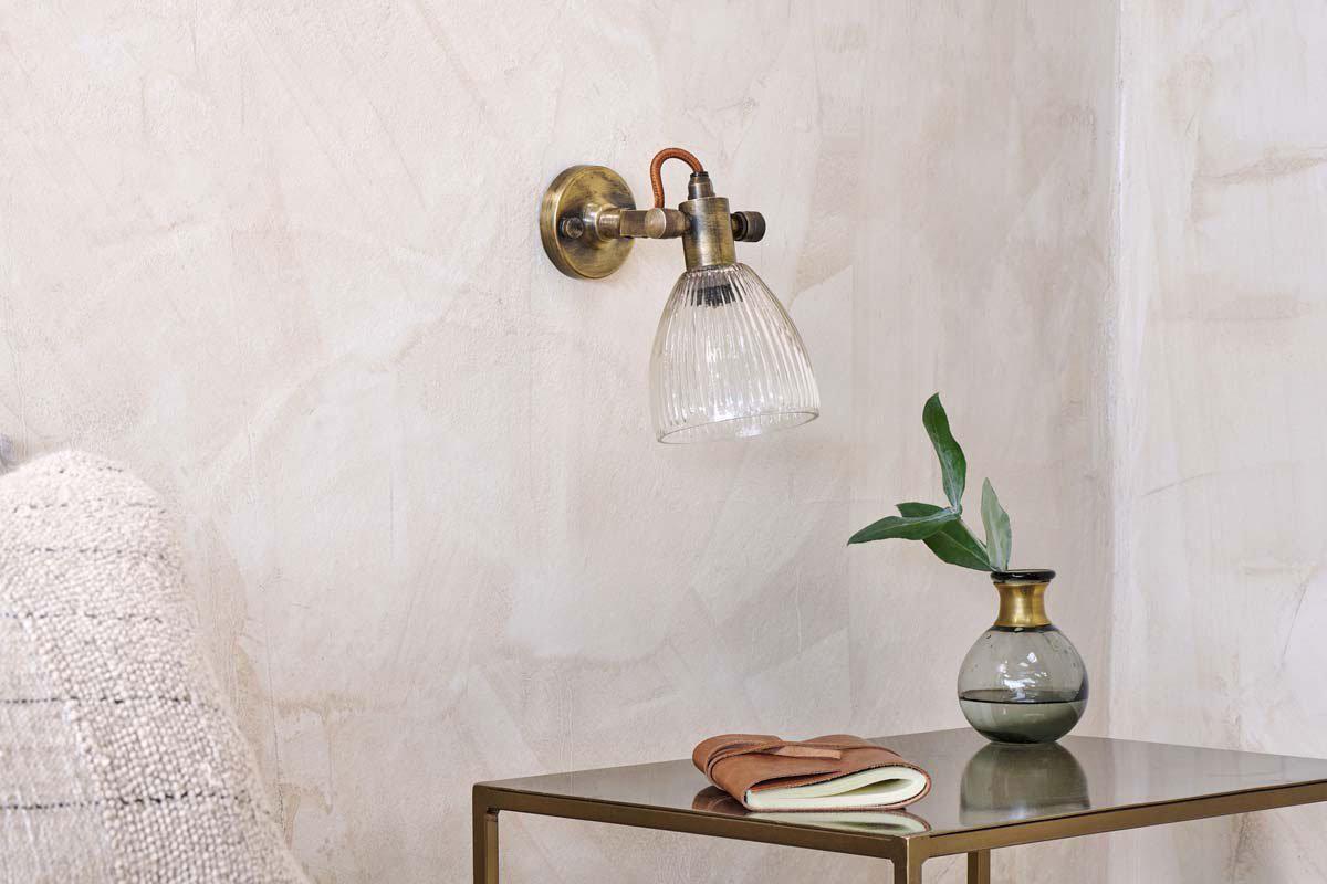 The nkuku Rarni Recycled Glass Single Wall Light is mounted on a textured beige wall above a metal side table with a green-leaf vase, closed notebook, and brown eyeglasses. The edge of a textured chair appears on the left.