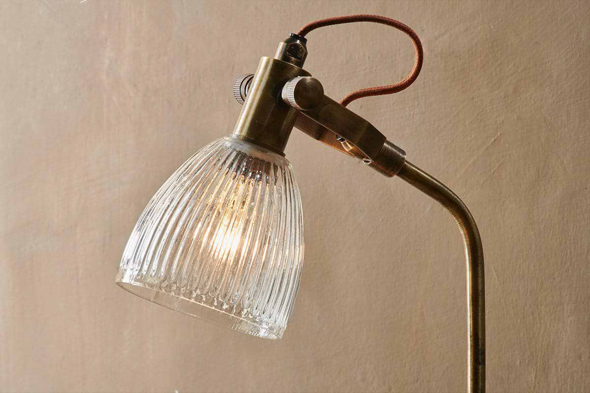 A close-up of the nkuku Rarni Recycled Glass Table Lamp in antique brass, featuring a ribbed clear glass shade, curved neck, exposed brown fabric cord, and warm light against a beige textured wall.