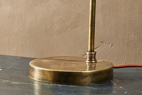 Close-up of the nkuku Rarni Recycled Glass Table Lamp in Antique Brass: a cylindrical stem and ribbed glass shade rest on a dark surface, with a brown fabric power cord extending toward a tan wall.