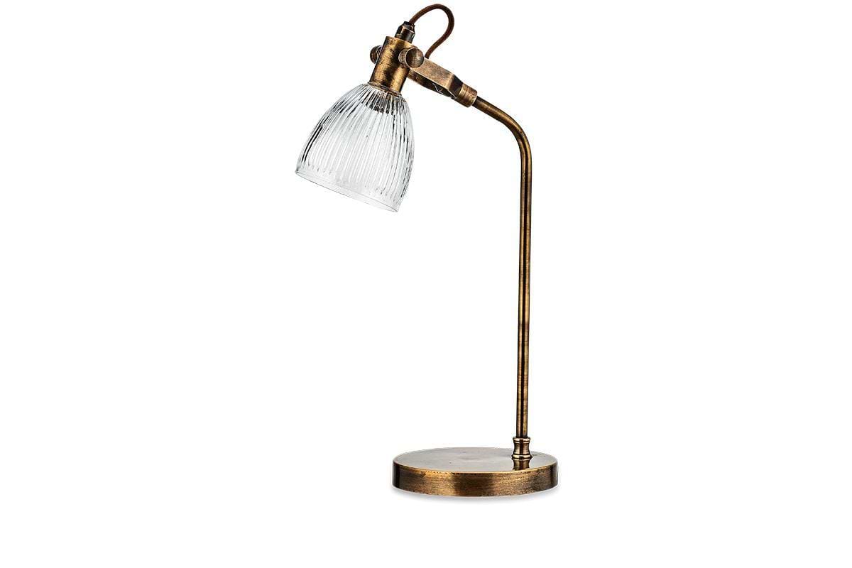 The nkuku Rarni Recycled Glass Table Lamp in Antique Brass features a curved arm, round base, and clear ribbed glass shade angled downward for an industrial vintage look. Shown against a white background.
