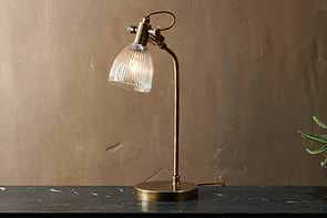 The nkuku Rarni Recycled Glass Table Lamp in Antique Brass, featuring a ribbed glass shade and curved arm, stands on dark marble. Its reddish-brown cord loops behind it against a plain brown wall, casting a warm light.