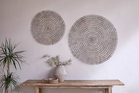 A minimalist interior showcases two large nkuku Rayan Wire Wall Art pieces in Antique Brass on a textured white wall, with a rustic wooden table below, accented by a striped ceramic vase, green branches, wooden bowl, and a potted spiky plant.