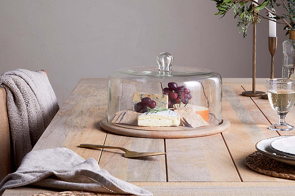 A cozy and elegant dining table is set with a cheese platter featuring assorted cheeses, crackers, and grapes displayed under the nkuku Recycled Glass Cake Dome. Surrounding the centerpiece are a wine glass, plates, a fork, napkins, and decorative greenery in the background.