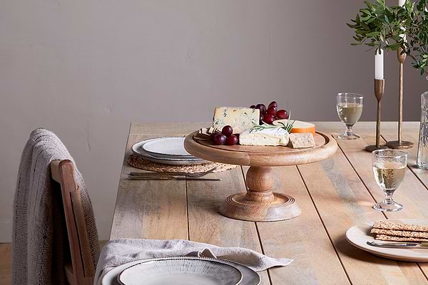 A wooden dining table for two with a mango wood base features beige chairs with gray cloth. At the center, cheese, grapes, and crackers rest under the nkuku Recycled Glass Dome Cake Stand, complemented by white wine glasses.