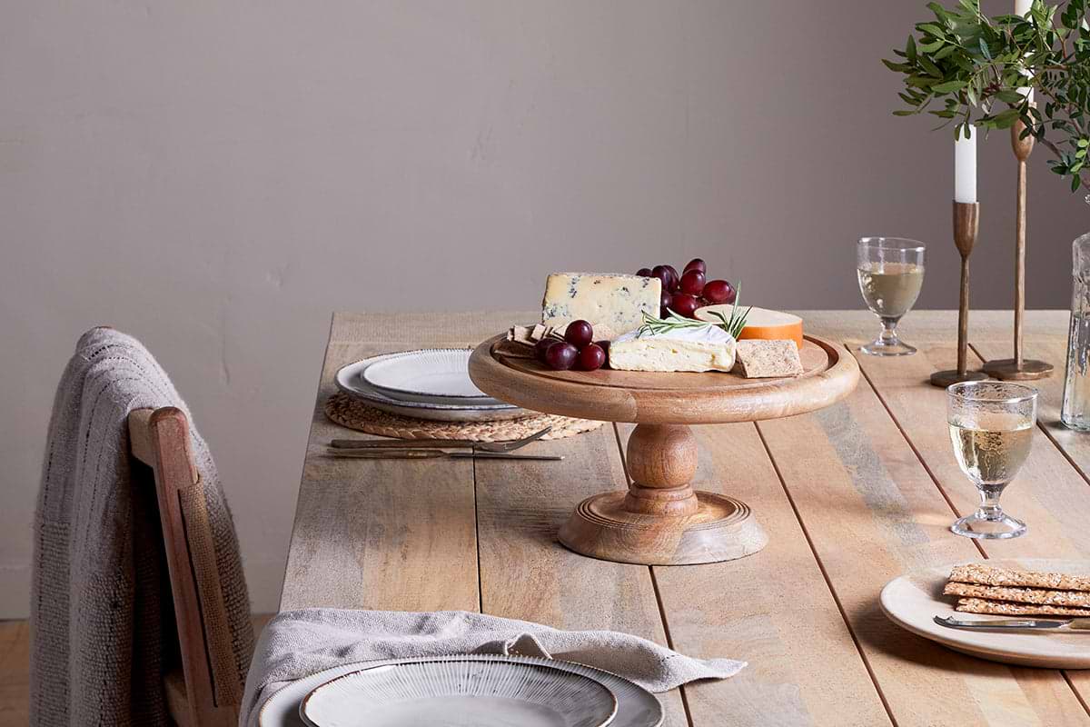 A wooden dining table for two with a mango wood base features beige chairs with gray cloth. At the center, cheese, grapes, and crackers rest under the nkuku Recycled Glass Dome Cake Stand, complemented by white wine glasses.