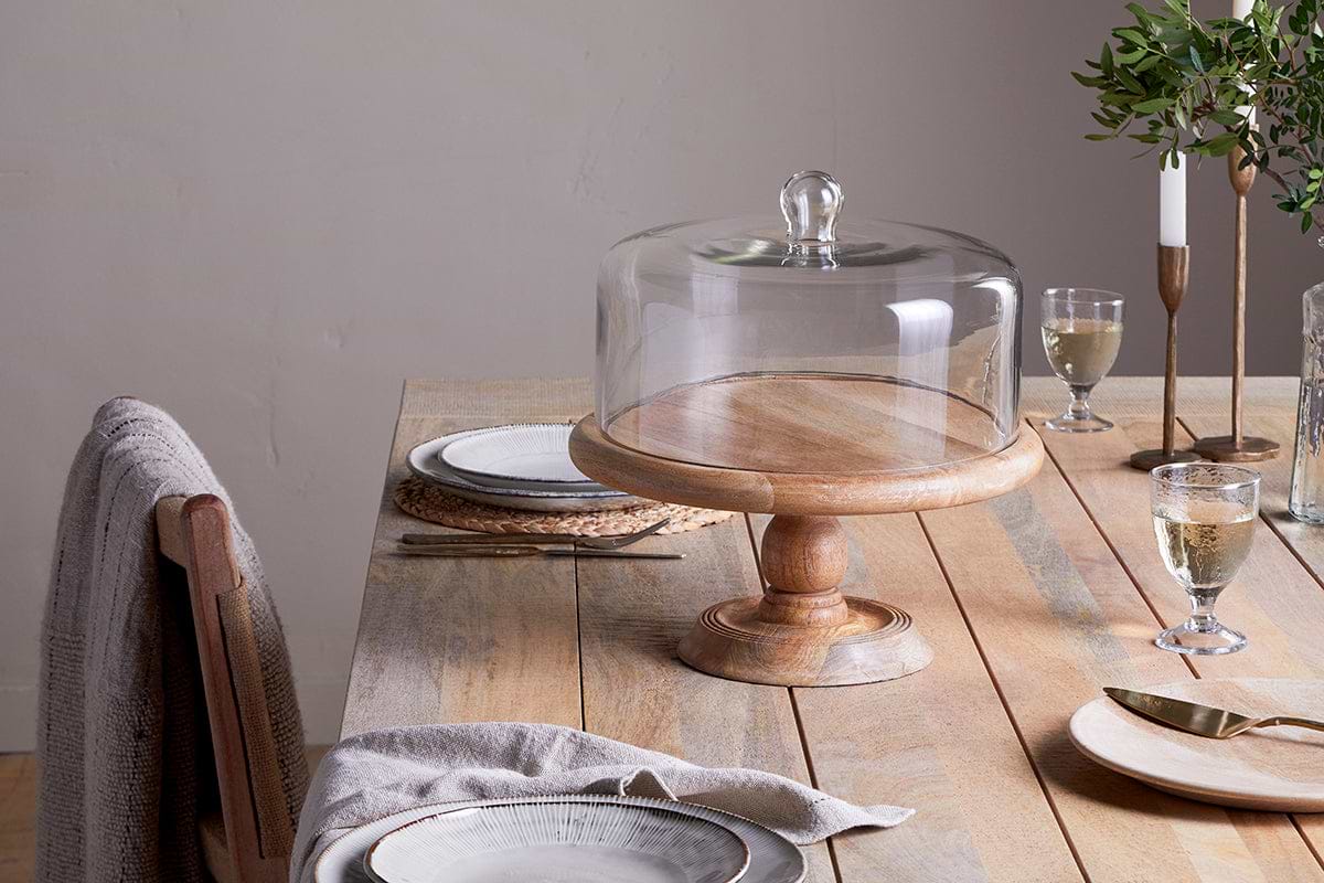 A rustic wooden table is set for dining with plates, cutlery, and white wine. At the center, nkuku’s Recycled Glass Dome Cake Stand with a mango wood base showcases desserts amid candles and greenery.