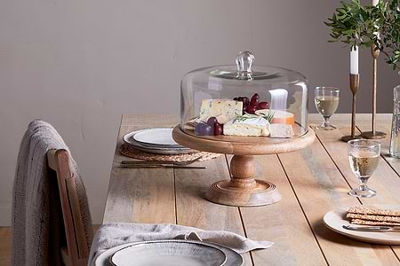 A rustic wooden table for two features white plates, woven placemats, and wine glasses. At the center, nkuku’s Recycled Glass Dome Cake Stand displays cheeses and grapes. Greenery and candlelight create a warm backdrop.