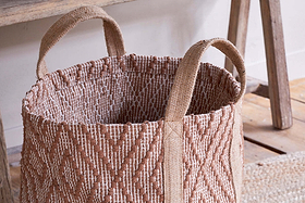 The Renu Jute Basket by nkuku features sturdy handles and a textured, geometric kilim design in rust and natural tones. It sits on a wooden floor beside rustic furniture in a cozy, neutral-colored room.
