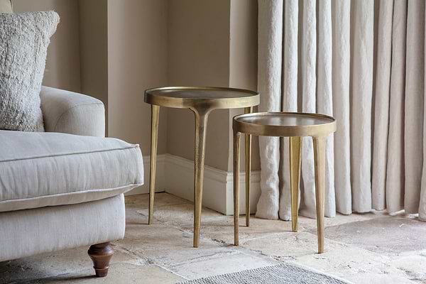 A pair of nkuku Rohia Cast Metal Side Tables in brass with fluted legs stand on a stone tile floor beside a beige upholstered sofa topped with a textured cushion, illuminated by soft natural light and pale curtains.