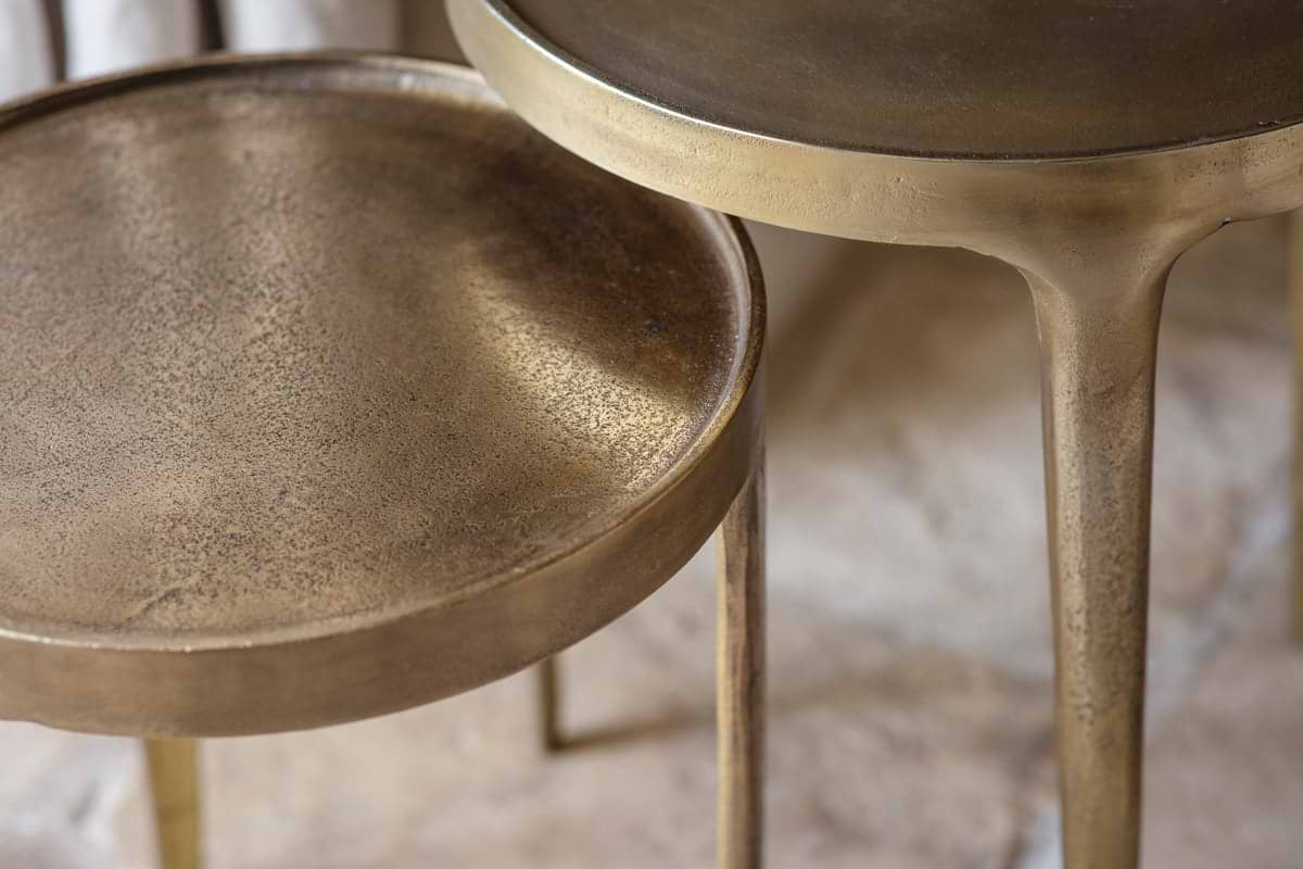Close-up of two nkuku Rohia Cast Metal Side Tables in brass with round tray tops and slender, fluted legs. The textured, antique gold finish displays a subtle dark patina. Smaller table is in front; larger one is partially visible behind.