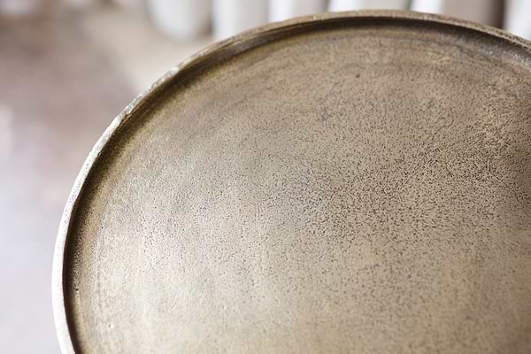 Close-up of the nkuku Rohia Cast Metal Side Table in brass, featuring a round aluminum frame with a textured, speckled gold surface and slightly raised edge. The blurred beige background suggests an indoor setting.