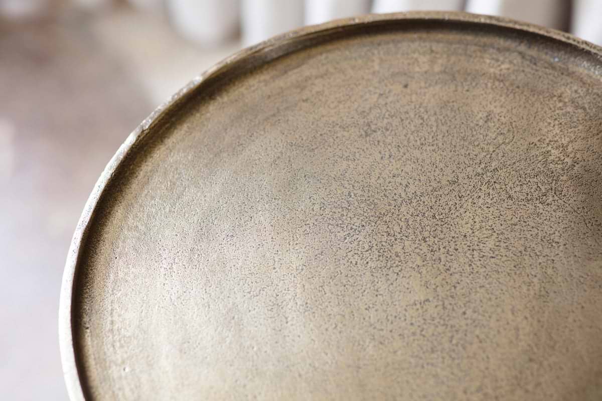 Close-up of the nkuku Rohia Cast Metal Side Table in brass, featuring a round aluminum frame with a textured, speckled gold surface and slightly raised edge. The blurred beige background suggests an indoor setting.