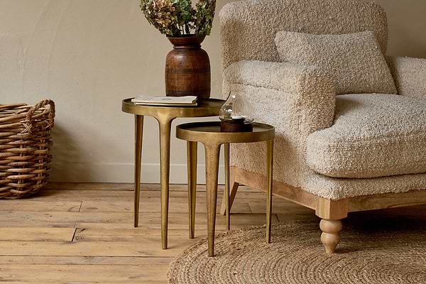 A cozy armchair with a textured cream cover sits beside two gold nesting tables, including the nkuku Rohia Cast Metal Side Table in brass. The tables display a wooden vase with dried flowers, a book, and glass ornament. A woven basket and rustic wood floor complete the scene.