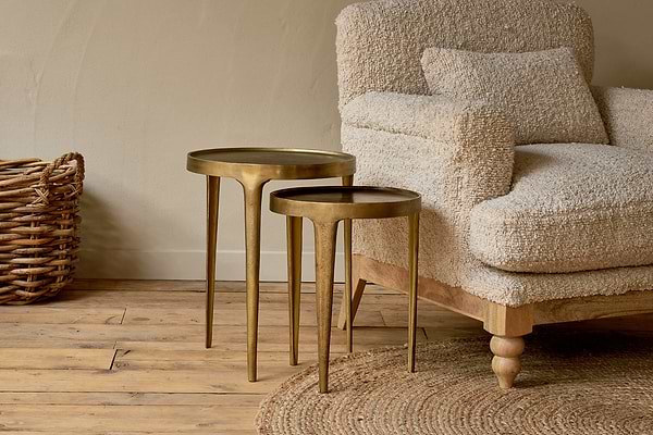 Two nkuku Rohia Cast Metal Side Tables in brass, featuring round tops and fluted legs, stand beside a beige armchair. A woven basket and circular jute rug complete the warm, rustic look on the wooden floor.