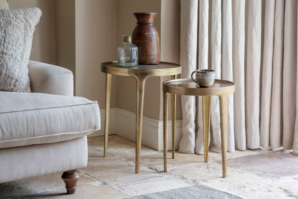 Two nkuku Rohia Cast Metal Side Tables in brass hold a vase and bottle on one, and a mug on the other. A neutral armchair sits to the left, framed by textured curtains behind.