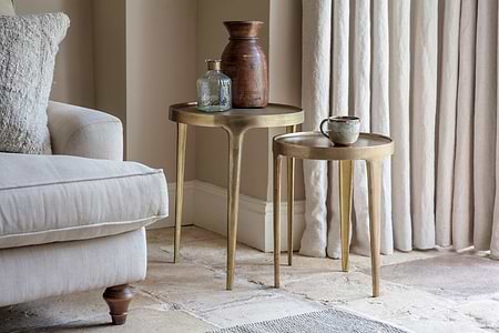 Two nkuku Rohia Cast Metal Side Tables in brass hold a vase and bottle on one, and a mug on the other. A neutral armchair sits to the left, framed by textured curtains behind.