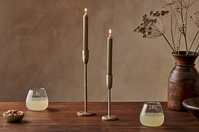 A wooden table with two glasses of a yellow drink, a small bowl of nuts, nkuku Rustic Soy Blend Dinner Candles in Olive Green (Set of 2) with textured brass holders, and a brown vase with dried flowers against a brown background.