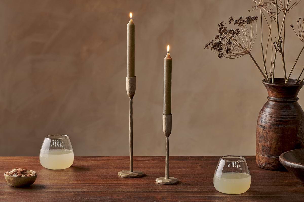 A wooden table with two glasses of a yellow drink, a small bowl of nuts, nkuku Rustic Soy Blend Dinner Candles in Olive Green (Set of 2) with textured brass holders, and a brown vase with dried flowers against a brown background.