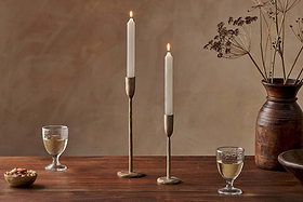A wooden table features two nkuku Rustic Soy Blend Dinner Candles in white, brass holders, two glasses of white wine, bowls of nuts and wood, and a tall brown vase with dried branches against a beige textured wall.