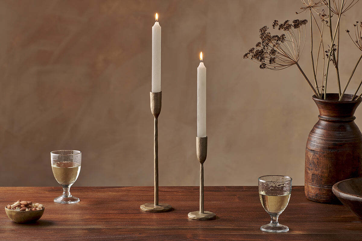 A wooden table features two nkuku Rustic Soy Blend Dinner Candles in white, brass holders, two glasses of white wine, bowls of nuts and wood, and a tall brown vase with dried branches against a beige textured wall.
