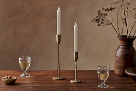 A wooden table displays two nkuku Rustic Soy Blend Dinner Candles in white, brass holders, wine glasses, a bowl of nuts, and a brown ceramic vase with dried flowers. A warm beige background adds to the cozy, elegant atmosphere.