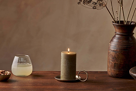 A lit Nkuku Rustic Soy Blend Pillar Candle in Olive Green (Small, textured finish) sits on a brass tray atop a wooden table, next to a clear glass of pale liquid, a bowl of nuts, and a tall wooden vase with dried flowers against a beige background.